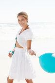 A blonde woman on a beach with a blue balloon wearing a white bolero and an embroidered skirt