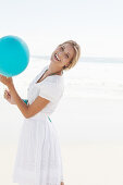 A blonde woman on a beach with a blue balloon wearing a white bolero and an embroidered skirt