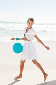 Reife blonde Frau mit blauem Luftballon in weißem Bolero und Rock mit Stickerei am Strand
