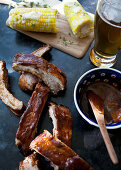 Pork ribs with barbeque sauce, a pint of beer and cobs of corn with thyme butter, on a black countertop