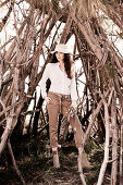 A dark-haired woman wearing a hat, a white shirt blouse and trouser in front of branches collected for a campfire