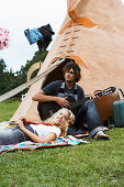 A woman lying on a bright blanket with a man with a guitar outside a teepee