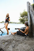 A young woman wearing a top and bikini bottoms on a river bank with a young man with a guitar sitting on the ground
