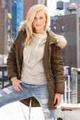 A young blonde woman wearing an olive-green jacket with a fur hood and jeans