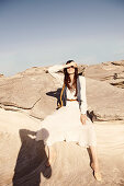 A brunette woman wearing a white blouse, a matching skirt and a gilet sitting on a rock