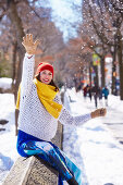A young woman wearing a red hat, a yellow scarf, a white jumper and blue trousers in a snowy park