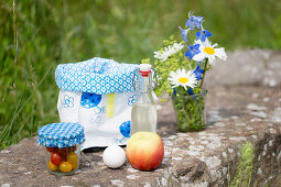 Posy in glass, oilcloth lunch bag, swing-top bottle, apple, egg and jar of tomatoes for picnic