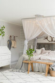 Double bed below canopy with fairy lights, wooden bench and bureau in pale bedroom