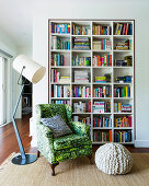 Antique armchair with green cover, pouf and floor lamp in front of bookshelf