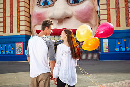 Junges Pärchen mit Ballons schaut sich in einem Freizeitpark vor einer Attraktion an