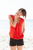 A mature brunette woman on a beach wearing a red shirt, a necklace and black shorts