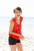 A mature brunette woman on a beach wearing a red shirt, a necklace and black shorts