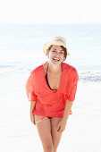 A mature brunette woman on a beach wearing a red shirt, a black bathing suit and a beige hat