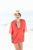 A mature brunette woman on a beach wearing a red shirt, a black bathing suit and a beige hat