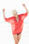 A mature brunette woman on a beach wearing a red shirt, a black bathing suit and a beige hat