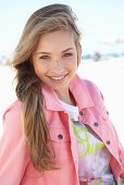 A young blonde woman on a beach wearing a printed t-shirt and a pink denim jacket