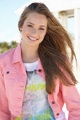 A young blonde woman on a beach wearing a printed t-shirt and a pink denim jacket