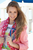 A young blonde woman on a beach wearing a printed t-shirt, a pink denim jacket and a patterned scarf