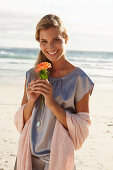 A mature blonde woman on a beach wearing a silver summer dress and holding an orange flower