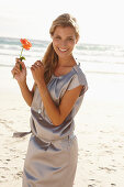 Reife blonde Frau mit silbernem Sommerkleid und orangener Blume am Strand