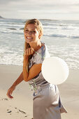 A mature blonde woman on a beach wearing a silver summer dress and holding a white balloon