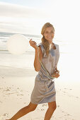 A mature blonde woman on a beach wearing a silver summer dress and holding a white balloon