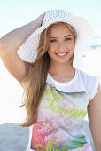 A young blonde woman on a beach wearing a colourful t-shirt and a white summer hat