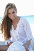 A young blonde woman on a beach wearing a white summer dress and a white summer hat