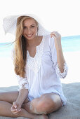 A young blonde woman on a beach wearing a white summer dress and a white summer hat