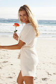 Reife blonde Frau in Dessous und Strickjacke und mit Blume in der Hand am Strand