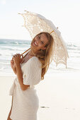 A mature blonde woman on a beach wearing lingerie and a cardigan with a parasol