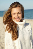 A young blonde woman on a beach wearing a white turtle neck jumper