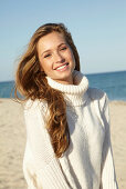 A young blonde woman on a beach wearing a white turtle neck jumper