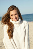 A young blonde woman on a beach wearing a white turtle neck jumper