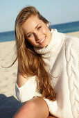 A young blonde woman on a beach wearing a white turtle neck jumper