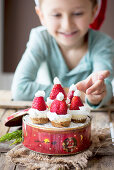 A boy sitting in front of mini cheesecakes in a biscuit tin decorated with strawberry Christmas hats