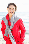 Brunette woman wearing read trench coat with grey sweater over shoulders on beach