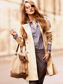 A young woman wearing grey trousers, a striped blouse, a beige trench coat with a bag