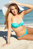 A young blonde woman on a beach wearing a turquoise bikini holding a white summer hat