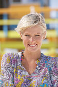 A mature blonde woman with short hair on a beach wearing a patterned summer dress