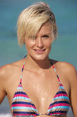 A mature blonde woman with short hair on a beach wearing a striped bikini