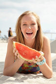 Junge blonde Frau im bunten Sommerkleid hält Melone am Strand