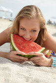 A young blonde woman on a beach wearing a colourful summer dress holding a wedge of melon