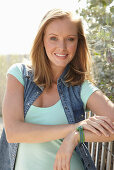 A young blonde woman outside wearing a sleeveless denim jacket and a light-blue top