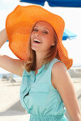 A young blonde woman on a beach wearing a light-blue summer dress and an orange summer hat