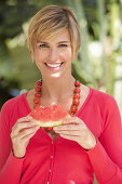 A mature woman with short blonde hair outside wearing a red shirt and holding a slice of watermelon