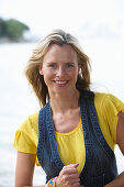 A young blonde woman on a beach wearing a yellow top and a denim waistcoat