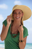A young blonde woman on a beach wearing a green top and a beige hat