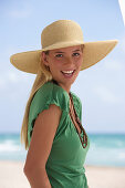 A young blonde woman on a beach wearing a green top and a beige hat