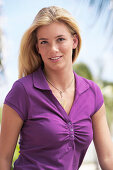 A young blonde woman under palm trees wearing a purple top